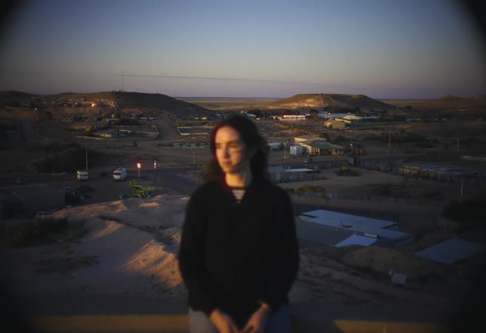 An out of focus woman sitting on a ledge looks out over a desert town at dusk while the sky transitions from orange to dark blue