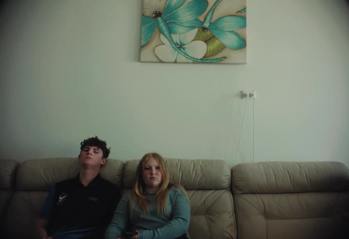 Two teenagers sit quietly on a couch beneath a butterfly painting their expressions subdued in a softly lit living room