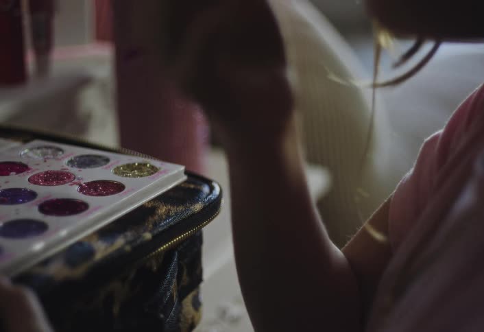 A close view shows a girl hand using a small brush with a colorful handle to dip into a palette filled with various shades of glittery makeup