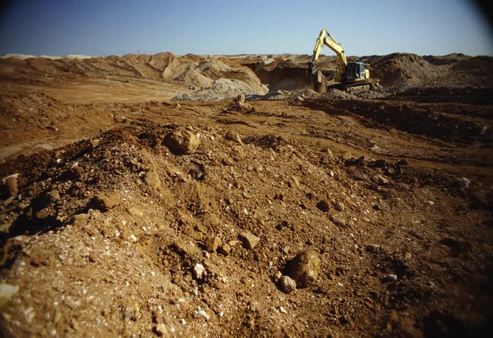 An excavator digs through dry earth at a large construction site under clear daylight