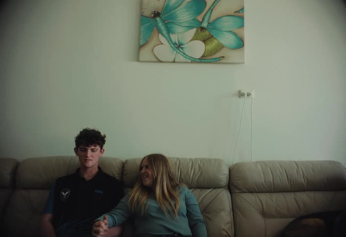 A young man and woman sit together on a creamcolored leather sofa smiling and looking at each other while holding hands in a brightly lit living room featuring a dragonfly painting on the wall above them