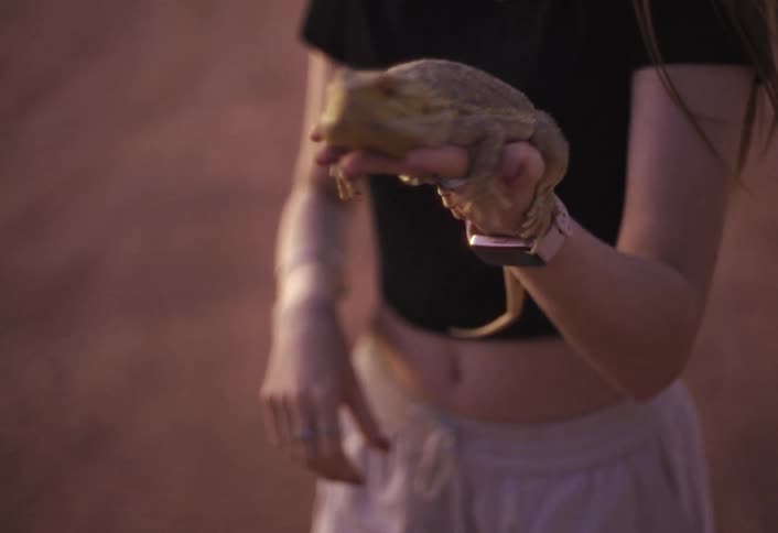 A young woman stands outdoors at midday holding a small lizard in her open palm
