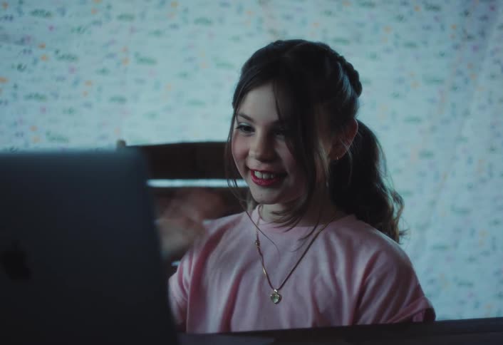 A young girl with braided hair and light makeup smiles and waves while looking at a laptop screen in front of patterned drapes