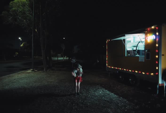 Bathed in the festive glow of a yellow food trucks colorful lights a woman in a red sequin dress and patterned jacket dances with freespirited energy on a dark roadside at night