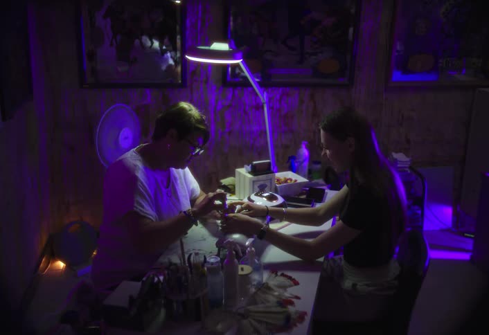In a dimly lit cluttered workshop with purple neon lighting a woman files the nails of a young girl seated at a nail station table