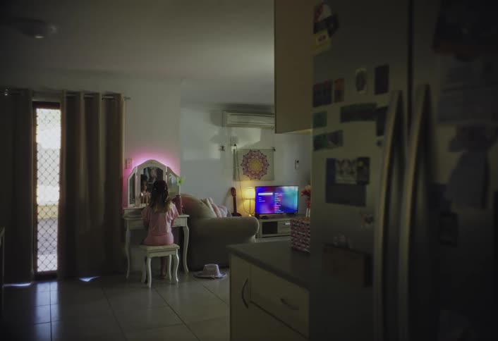 A young girl sits at a vanity with a glowing mirror in a cozy living space while a television flickers softly in the background