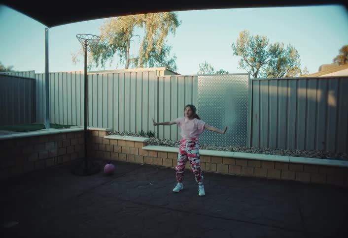 A young girl in a pink shirt and floral pants performs a dance routine on a paved backyard patio near a basketball hoop and a metal fence