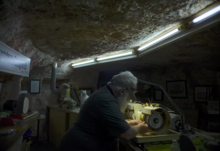 An older man works intently at a grinding machine in a dim cavelike workshop illuminated by fluorescent lights