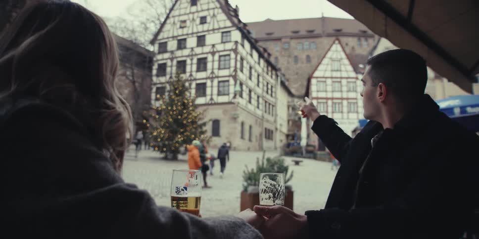 A couple sits at an outdoor bar at midday each with a glass of beer watching the street while they talk