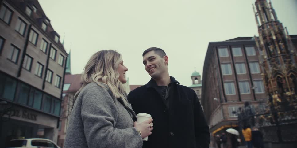 On a hazy day in a historical city a couple walk arm and arm talking and smiling while sightseers are seen in the background