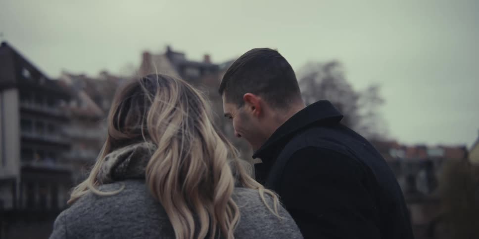 A happy couple walks together along a bridge or waterfront sharing smiles and intimate glances the man keeps a supportive arm around the womans waist as they enjoy a cold overcast day in a european city