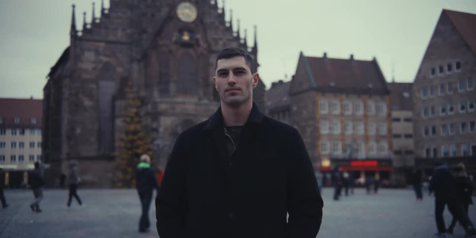 A man in a black coat stands in a public square in front of a large gothic church smiling and looking around while people walk in the blurred background