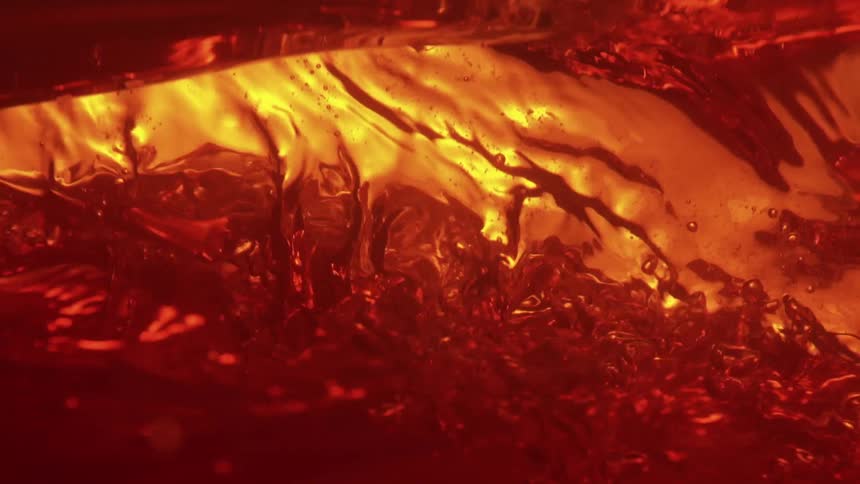 A close view shows golden ripples and bubbles forming in a dark red liquid as it is stirred or poured