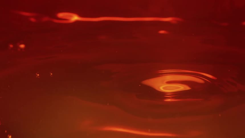 An orange liquid surface experiences a heavy drip that creates a deep glowing crater followed by a rising rebound ripples then spread outward across the dark red viscous fluid as the surface gradually settles back into a steady flow