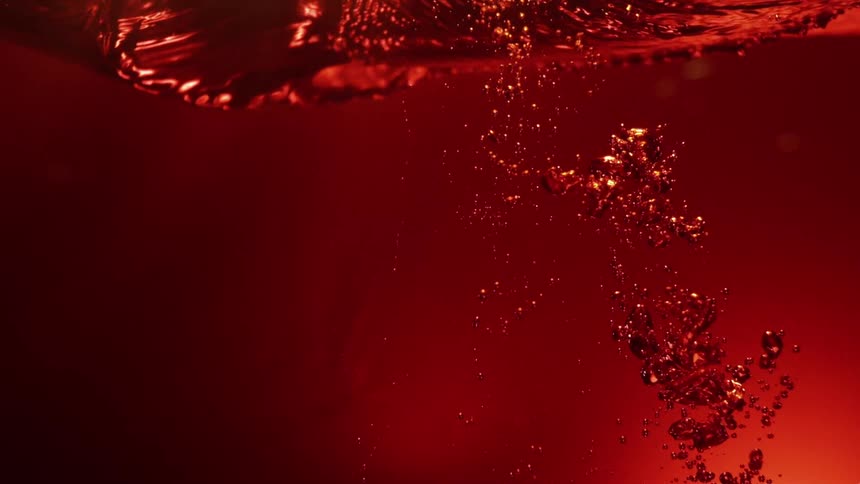 A close view shows air bubbles rising and swirling through a deep red liquid under a bright light