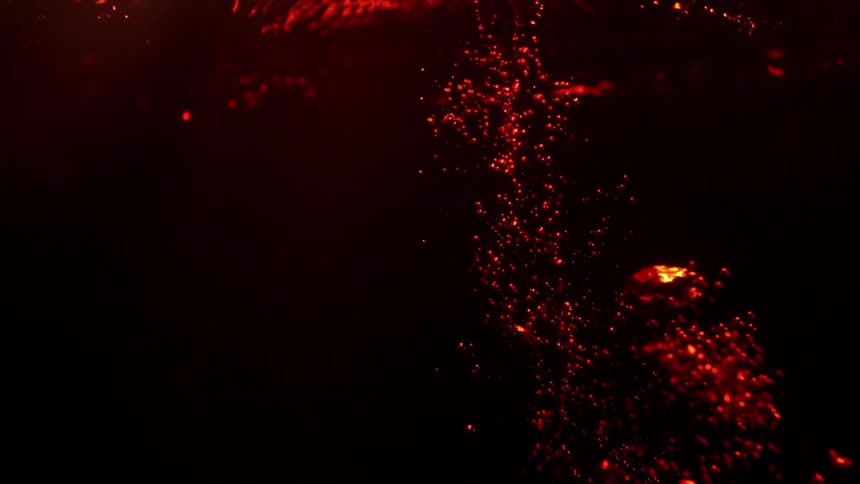 Red liquid pours into a dark container from above creating a turbulent stream of rising bubbles and swirling amber currents against a black background