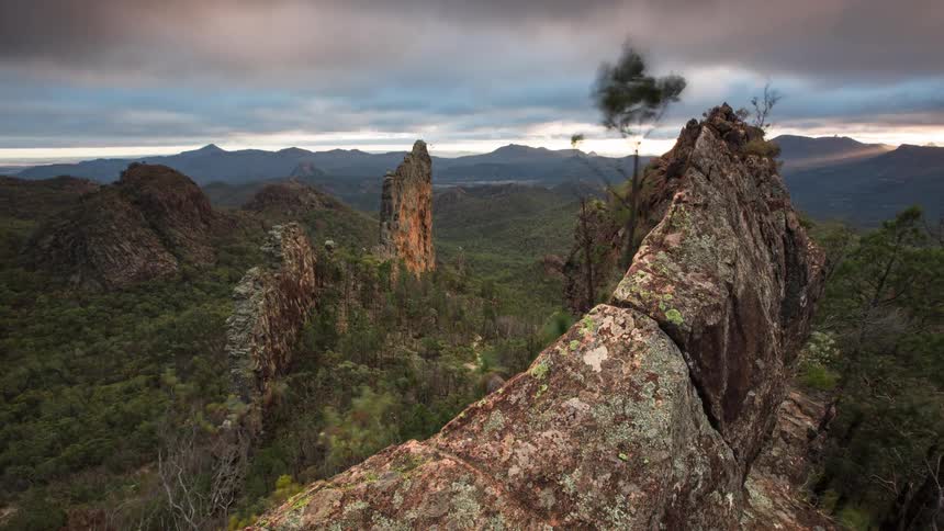 A wide shot captures a jagged rock formation and a prominent stone pinnacle standing amidst a vast mountain range under a heavy overcast sky