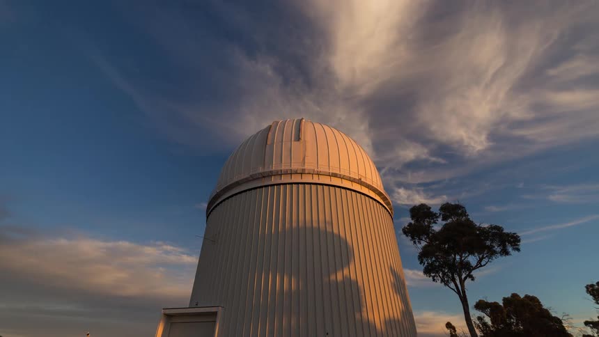 A white astronomical observatory dome stands tall against a vast open sky clouds drift rapidly overhead as the light shifts from bright daylight to the warm hues of sunset
