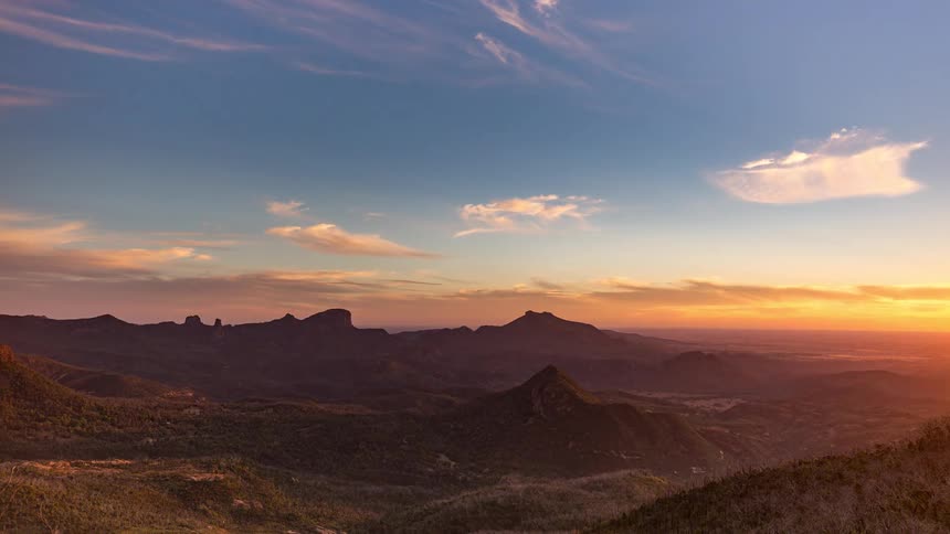 A wideangle timelapse captures the transition from day to dusk over a rugged mountain range showing wispy clouds moving across a blue and orange sky as shadows deepen across the rocky peaks and valleys of the vast natural landscape