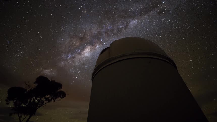 A timelapse captures the milky way and a vast field of stars rotating over a large observatory dome and a dark tree silhouette at night