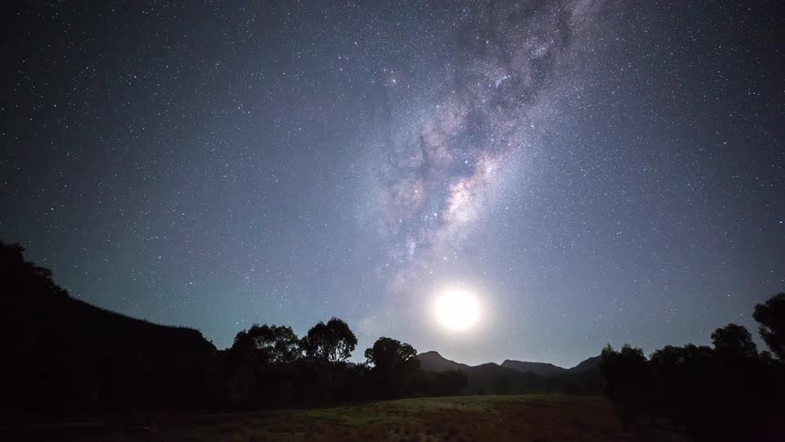 Stars of the milky way move over trees on mountains in the night sky and fade with the rising sun
