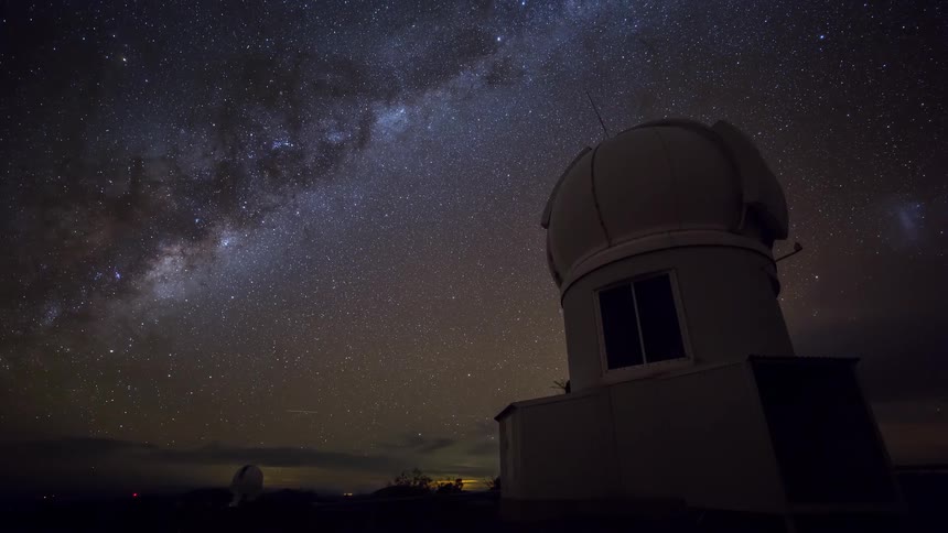 A timelapse captures the milky way galaxy moving across a starfilled night sky behind the silhouette of a white astronomical observatory dome while thin clouds pass over the dark horizon