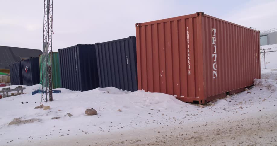 Several shipping containers in various colors stand in a row on a snowy gravelcovered industrial lot under a gray sky with a metal utility tower visible in the background