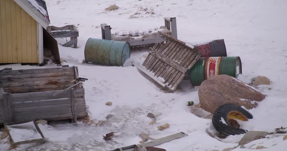 Various metal barrels and wooden crates lie scattered across a snowy trashstrewn ground next to a yellow building