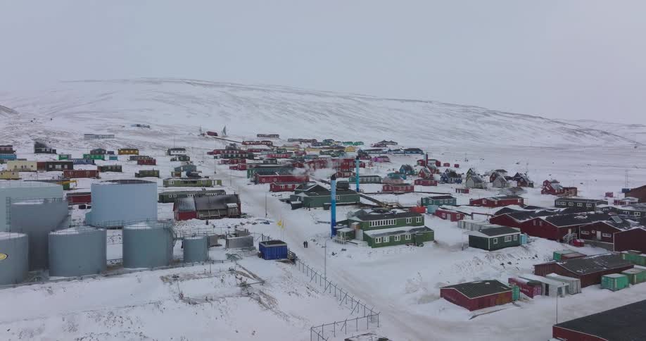 A sweeping view drifts over large industrial tanks to reveal an isolated settlement of vibrant red green and blue houses perched on a vast snowcovered mountain slope