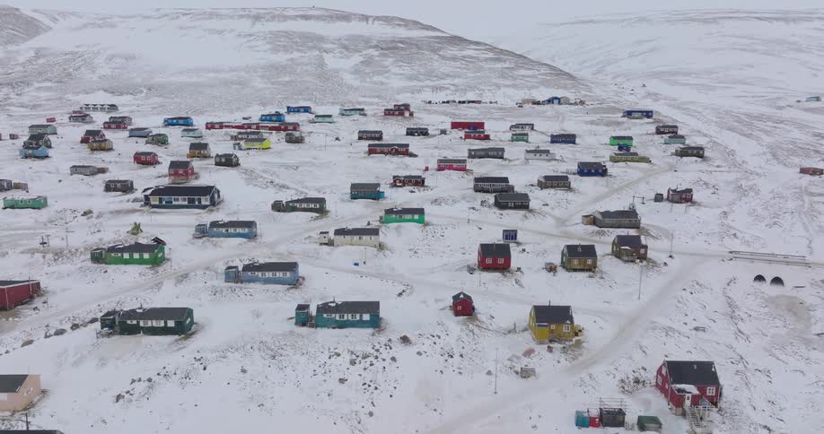 An aerial shot shows a remote arctic village with numerous colorful houses scattered across a snowy hillside under an overcast sky with small roads and utility lines connecting the various small rectangular buildings in the winter landscape