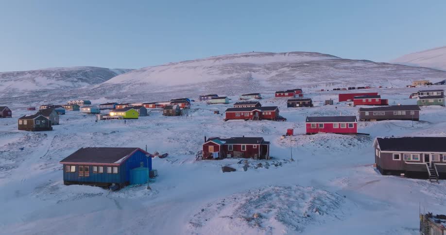 A small town sits on a snowy hillside at midday with buildings scattered across the slope and winter light reflecting off the white landscape