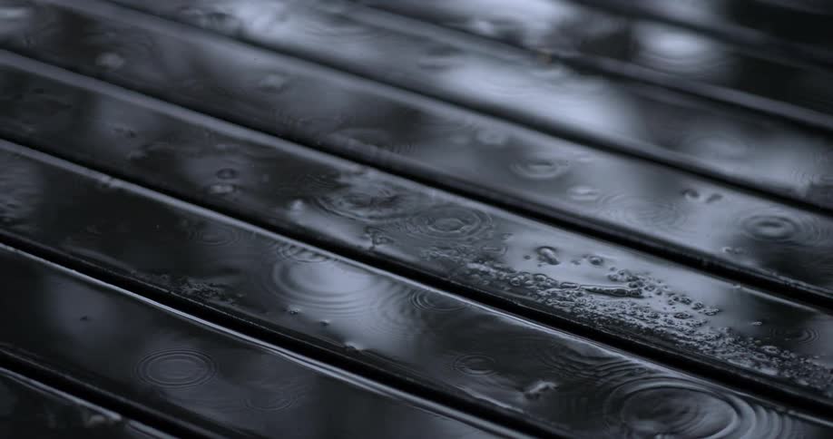 Raindrops fall and create ripples on a dark wet wooden surface with horizontal slats