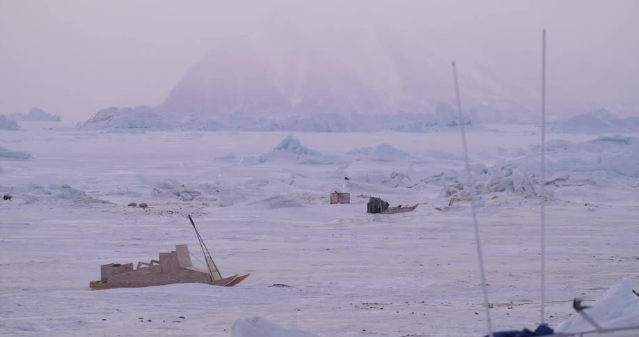 Traditional wooden sleds rest upon the endless frozen ice sheet dwarfed by a massive fogshrouded mountain rising into the soft pinkhued sky