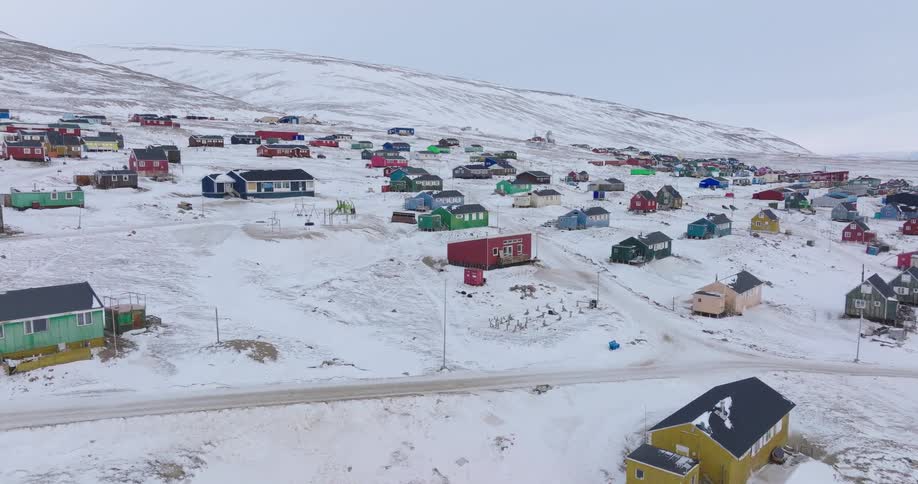 An aerial drone shot glides over a remote arctic village built on a snowy hillside showcasing dozens of small brightly colored houses in shades of red blue and green connected by dirt roads under a vast overcast winter sky