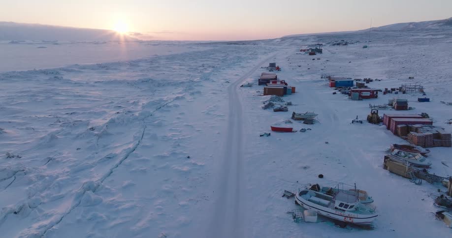 Road in a snowcovered polar region with boats and scattered structures along the route at sunset