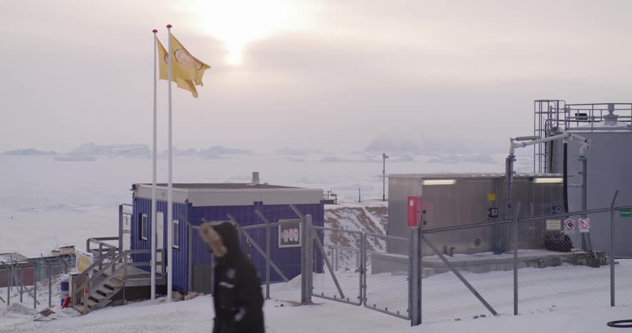 People walk past a building and machinery inside a fence along the arctic coast