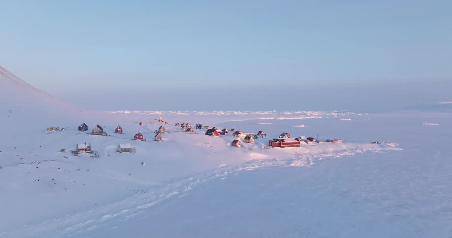 An aerial drone shot captures a cluster of colorful houses in a remote arctic village nestled on a snowcovered hillside overlooking a frozen coastline under the soft pale light of a clear winter sky