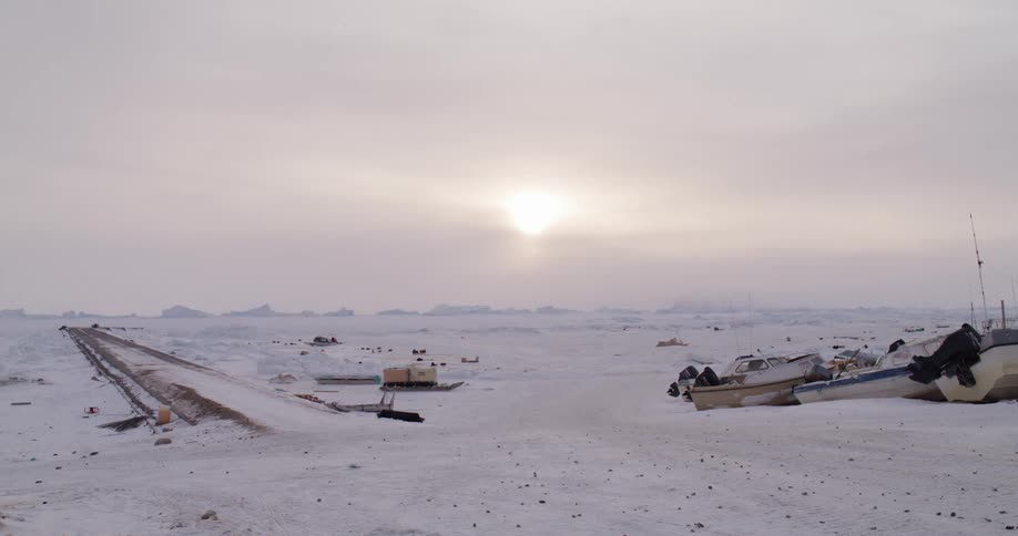 Dog sleds move across the snowy terrain in the distance while boats lie on snow covered land near a paved surface on a winter day
