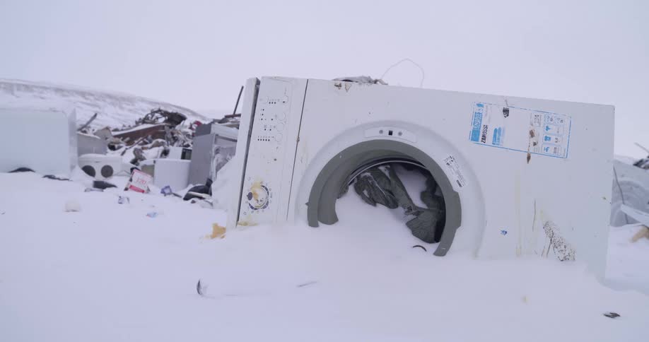 Broken washing machine on the snow surrounded by garbage outdoors during day time