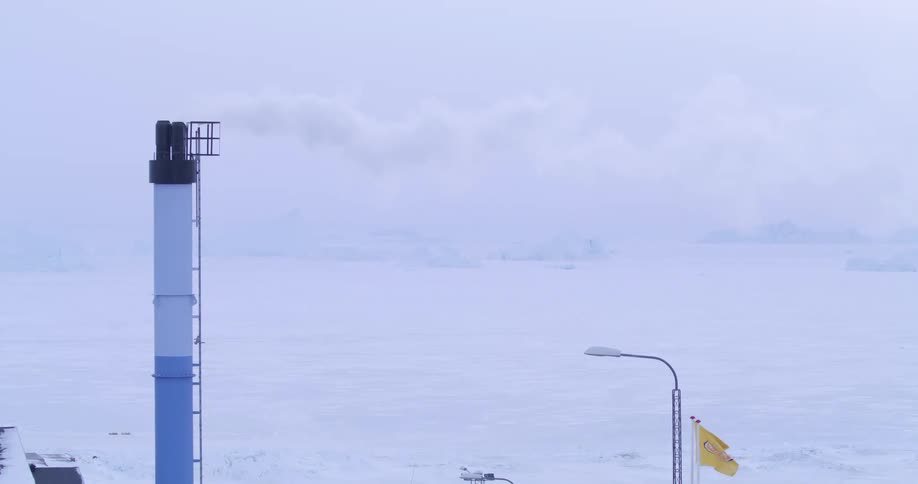 Smoke rises from a tall blue and white industrial chimney overlooking a frozen arctic bay where a yellow flag waves in the wind near small coastal buildings and distant icebergs under a cloudy sky