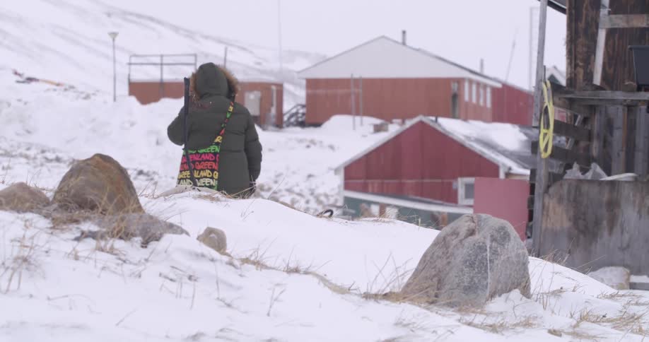 A person wearing a heavy parka and a colorful bag walks away through a snowy landscape past red village houses and rocky terrain on a cold overcast winter day