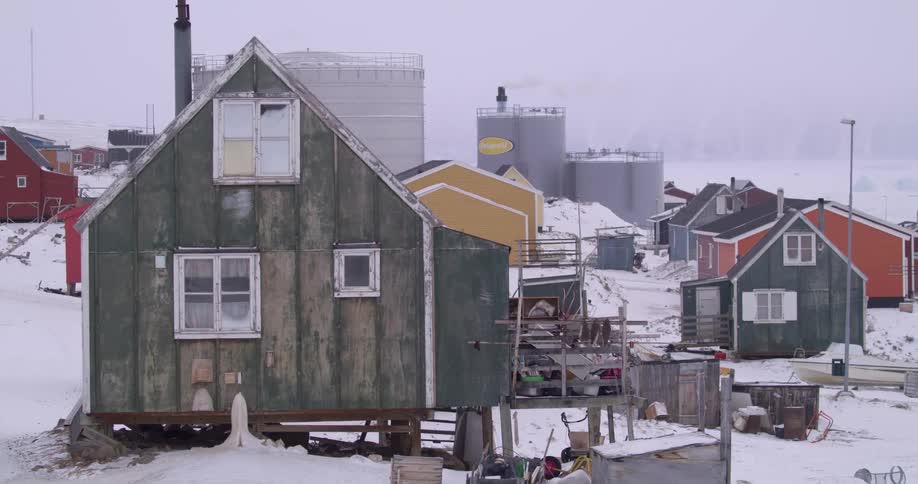 Various colorful houses sit on a snowy hillside in a remote village with large industrial storage tanks rising behind the residential structures under a hazy and overcast winter sky