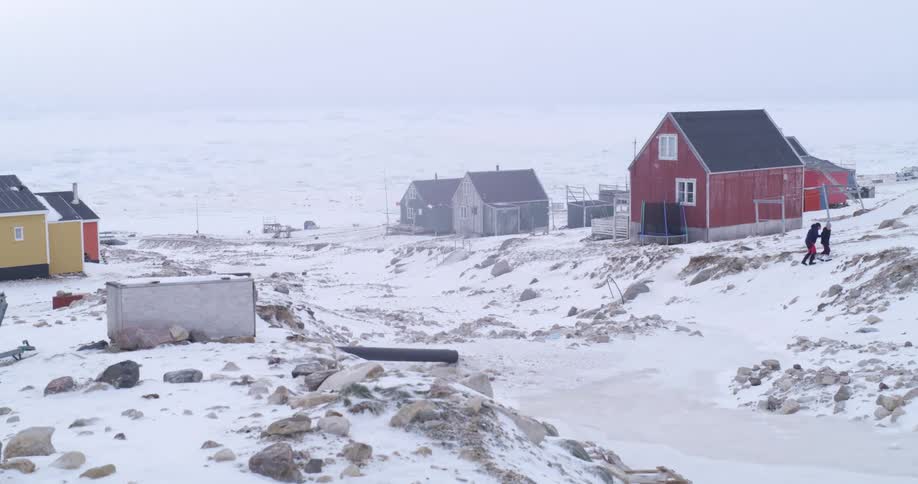 Two young people climb up a snow and ice covered hill near buildings in the arctic