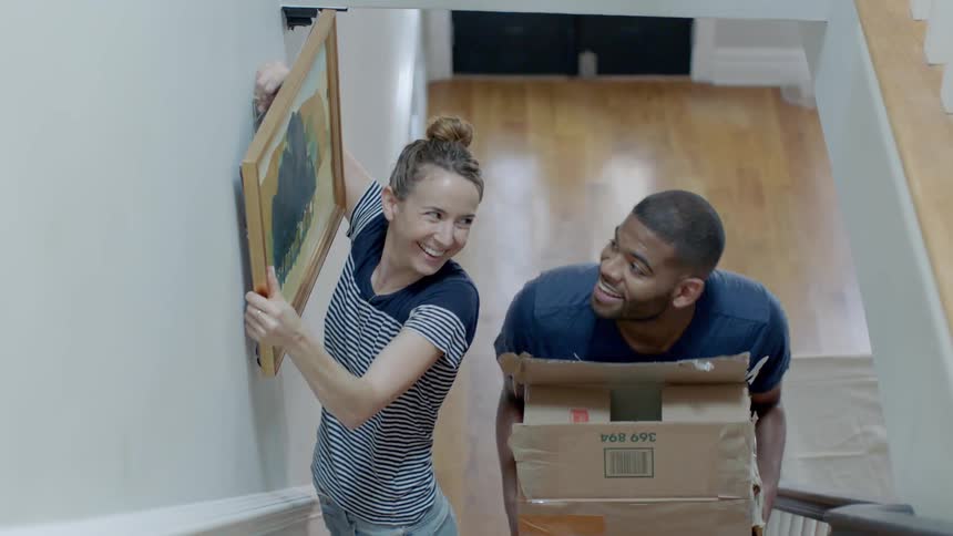 A woman hangs a picture in a stairwell while a man carries a cardboard box upstairs