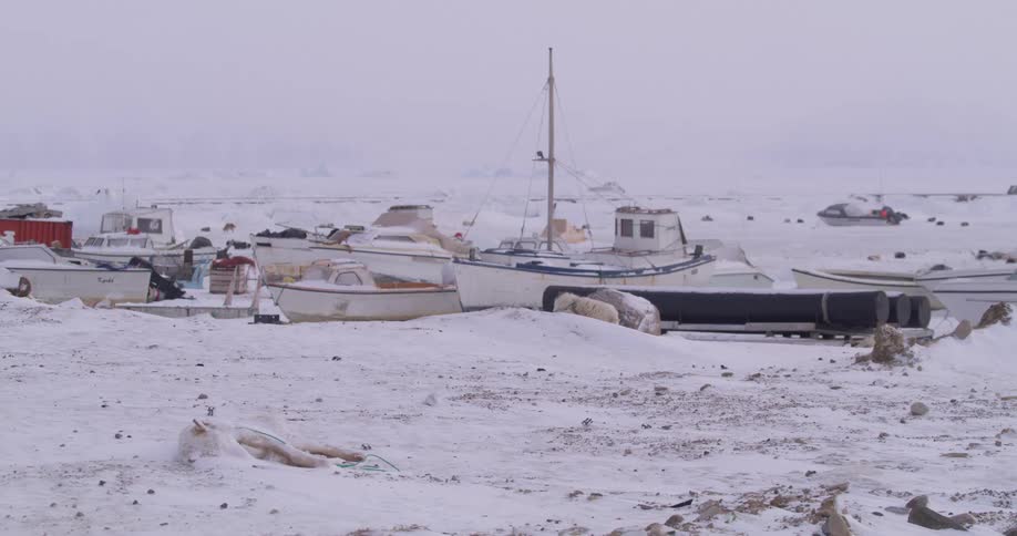 Several small boats and a large black pipe sit on a snowy desolate shore under a grey foggy sky