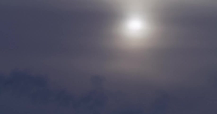 A distant view shows the sun shining through a layer of thick grey clouds in a hazy sky