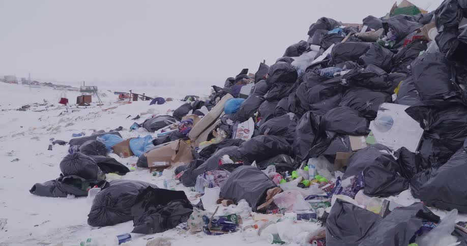 A large pile of black trash bags and loose garbage sits in a landfill covered in snow under a grey sky showing various types of plastic waste and cardboard containers scattered across the frozen ground in a wide shot of the dump