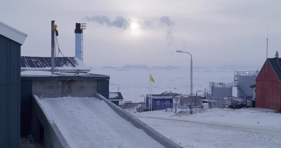 Industrial infrastructure in a polar settlement with a smokestack emitting smoke and a frozen sea in the background