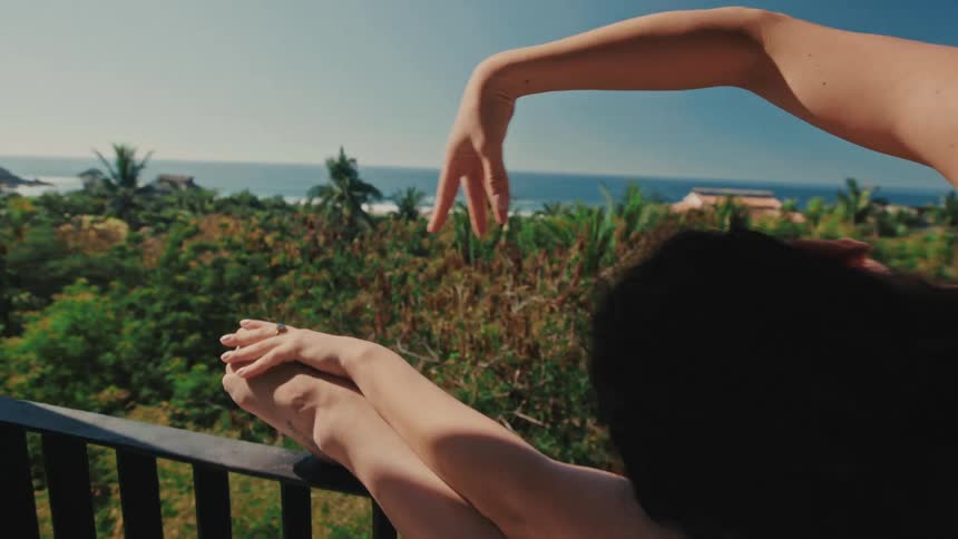 Viewed from behind a woman stretches on a balcony with her leg resting on the railing her hand reaching gracefully toward her foot against a backdrop of tropical greenery and the sea