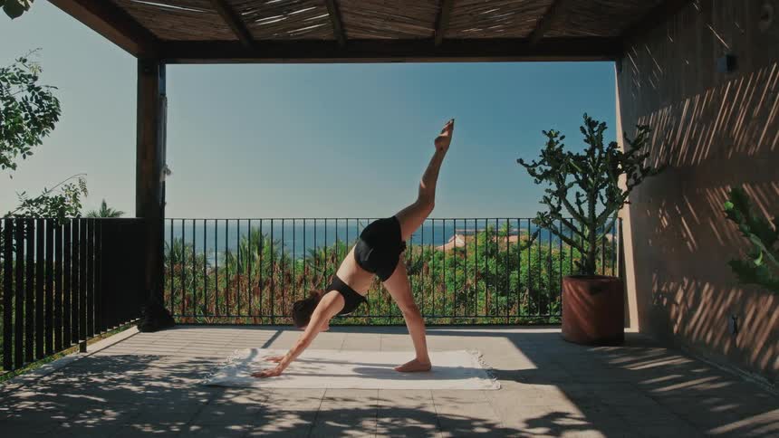A young woman stands on her front porch at midday dressed in activewear as she moves through a series of yoga stretches at midday